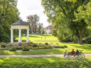 Eine grüne Parklandschaft mit einem eleganten Pavillon und zwei Personen, die auf einer Bank sitzen und die Aussicht bewundern.