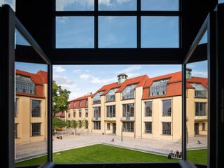 Blick auf das Hauptgebäude der Bauhaus-Universität Weimar, Foto: Thomas Müller, weimar GmbH Durch ein offenes Fenster sieht man ein modernes Gebäude mit roten Dächern und großen Fenstern, umgeben von Grünflächen.