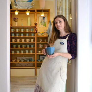 Keraminka Ute Raabe Die Frau mit langen Haaren und einem schlichten Kleid mit Schürze steht an einer Tür und präsentiert stolz eine blaue Tasse vor einem Geschirrregal.