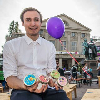 Bierbereis Felix Bieber Der Mann sitzt auf einer Holzbank vor dem Theater und präsentiert stolz die Eissorten von Biebereis.