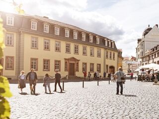 Goethes Wohnhaus, Foto: Jens Hauspurg, weimar GmbH Eine belebte Straße mit Menschen, die an einem gelben Gebäude vorbeigehen, während die Sonne scheint und Wolken am Himmel sind.