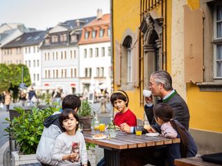 Brotklappe Eine Gruppe von Kindern und einem Erwachsenen genießt ein Frühstück im Freien, umgeben von bunten Gebäuden und Pflanzen.