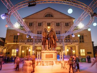 Weimarer Weihnacht, Foto: Marcus Glahn, weimar GmbH Eine festliche Szenerie mit einer Statue von Goethe und Schiller, umgeben von Lichtern und fröhlichen Menschen auf einer Eisbahn.