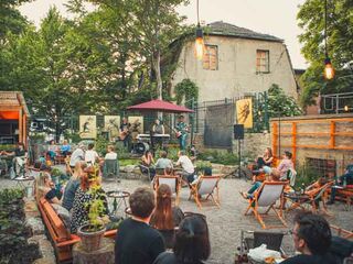Künstlergarten In einem gemütlichen Außenbereich genießen Gäste die Musik einer Band, die auf einer Bühne mit Pflanzen im Hintergrund spielt.