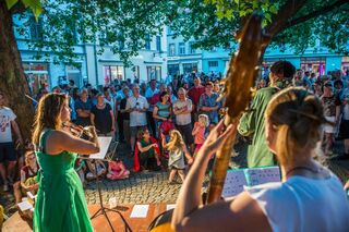 Fete de la musique Eine Musikerin in einem grünen Kleid spielt Geige vor einer großen Menschenmenge unter einem Baum in der Abenddämmerung.