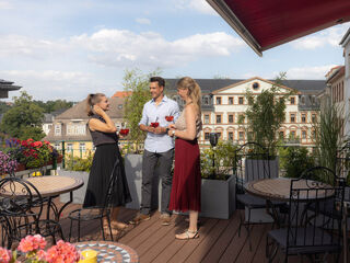 Auf einer stilvollen Terrasse stehen zwei Frauen und ein Mann, die mit Cocktails anstoßen und die Aussicht auf die Stadt genießen.