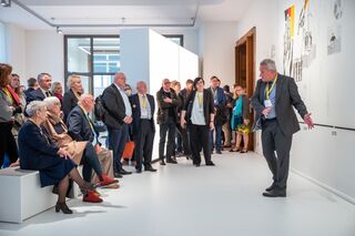 Prof. Dr. Jens Wagner erklärt den letzten Ausstellungsraum, Foto: Thomas Müller, Museum Zwangsarbeit im Nationalsozialismus Eine Gruppe von Menschen hört einem Redner in einem modernen Ausstellungsraum aufmerksam zu, während sie an einer Wand mit Kunstwerken stehen.