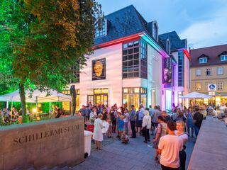 Schiller Museum Eine lebhafte Szene vor dem Schiller-Museum, wo Besucher in geselliger Runde unter bunten Lichtern und Zelten feiern.
