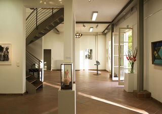 Foto: Galerie Eigenheim Ein heller Ausstellungsraum mit einer geschwungenen Treppe, Kunstwerken an den Wänden und einer offenen Tür zum Garten.