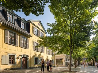 Schillers Wohnhaus Die Straße ist von Bäumen gesäumt, während Passanten an einem malerischen gelben Haus vorbeigehen und die Sonne genießen.