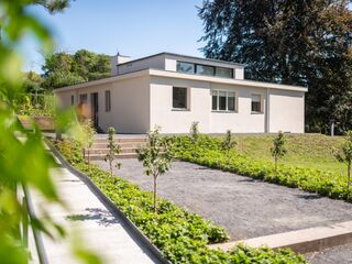 Haus am Horn Ein modernes, helles Haus mit großen Fenstern steht in einer grünen Umgebung, umgeben von kleinen Bäumen und Sträuchern.