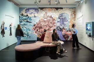 Ausstellungsimpression Faust. Eine Ausstellung In einem Ausstellungsraum sitzen zwei Besucher auf einer geschwungenen Bank, während sie Kopfhörer tragen und auf eine Wandzeichnung blicken.