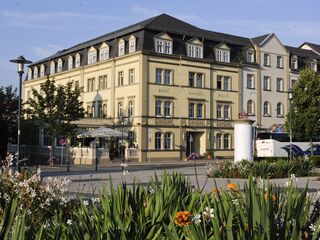 Außenaufnahme, Foto: Kaiserin Augusta Ein elegantes Hotelgebäude mit gelber Fassade, umgeben von blühenden Blumen und Bäumen, strahlt eine einladende Atmosphäre aus.