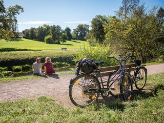 Schlosspark Tiefurt, Foto: Thomas Müller Zwei Radfahrer sitzen am Ufer der Ilm im Schlosspark Tiefurt. Die Fahrräder wurden an einer Sitzbank angelehnt.