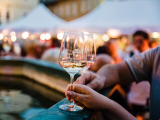 Weinfest Im Vordergrund sind zwei Gläser mit Weißwein zu sehen, umgeben von einer lebhaften Menschenmenge und festlichen Ständen.