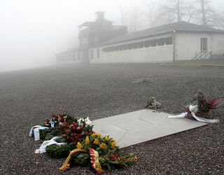 Gedenkstein Appellplatz, Foto: Gedenkstätte Buchenwald Ein stiller, nebliger Platz mit einem Gedenkstein und Blumen, der die Erinnerung an die Vergangenheit ehrt, im Hintergrund ein Gebäude.