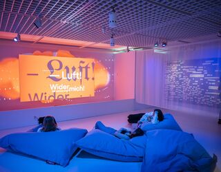 Ausstellungsimpression Faust. Eine Ausstellung In einem modernen Raum liegen mehrere Personen auf großen blauen Kissen und schauen auf eine projizierte, farbenfrohe Textbotschaft.