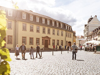 Goethe Nationalmuseum Ein sonniger Tag in einer Stadt, wo Passanten an einem Brunnen vorbeigehen und Cafés im Hintergrund sichtbar sind.