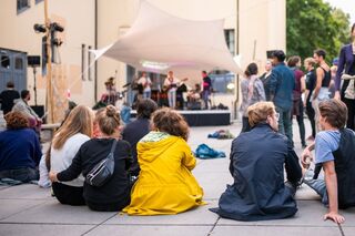 Studierende waehrend der summaery an der Bauhaus-Universitaet Weimar Eine Gruppe von Studierenden sitzt auf dem Boden und schaut auf eine Live-Band, die auf einer kleinen Bühne während der summaery spielt.
