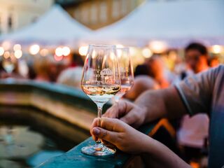 Weinfest Im Vordergrund sind zwei Gläser mit Weißwein zu sehen, umgeben von einer lebhaften Menschenmenge und festlichen Ständen.