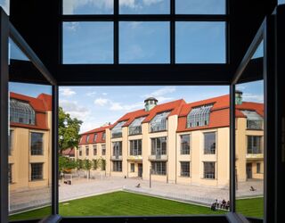 Blick auf das Hauptgebäude der Bauhaus-Universität Weimar Durch ein offenes Fenster sieht man ein modernes Gebäude mit roten Dächern und großen Fenstern, umgeben von Grünflächen.