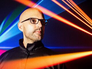 Ein Mann im Fokus mit bunten Lichtstrahlen im Hintergrund Ein Mann mit Glatze und Brille steht vor einem bunten Lichtspiel aus roten, blauen und grünen Strahlen.