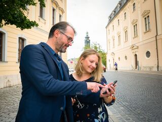 Paar in Weimar Ein Mann und eine Frau stehen auf einer gepflasterten Straße und schauen gemeinsam auf ein Smartphone, lächelnd und interessiert. im Hintergrund ist der Schlossturm zu sehen.