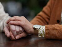 Eine Person greift die Hand einer weiteren Version, die linke Hand liegt schützend oben auf der Hand der zweiten Person.