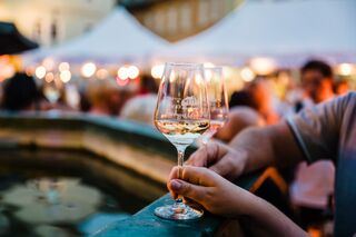 Weinfest Im Vordergrund sind zwei Gläser mit Weißwein zu sehen, umgeben von einer lebhaften Menschenmenge und festlichen Ständen.